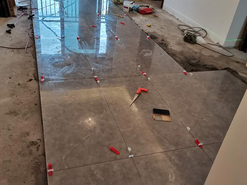 客廳地磚鋪貼：地磚鋪貼前，應(yīng)將地面清掃干凈，尤其是凝結(jié)后的混凝土，否則，地磚鋪貼后會出現(xiàn)翹邊、空鼓等現(xiàn)象，然后再灑水潤濕地面。地面充分濕潤，但不能有積水，然后將3:1混合好的沙子水泥鋪在地面上，進行基層處理。在地磚背面均勻抹水泥砂漿作為粘合層，厚度不低于1cm，然后鋪在剛才試鋪的位置上，用橡皮錘輕敲地磚飾面直至密實平整達到要求。地磚鋪完后24h進行清理勾縫，勾縫前應(yīng)先將地磚縫隙內(nèi)雜質(zhì)擦凈，用專用填縫劑勾縫，完工后（一般宜在24h之后）再用棉紗等物對地磚表面進行清理。